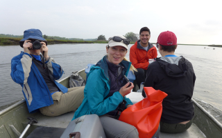 four people on a small row boat on the water. one on left has a camera to face. one in back has a red jacket. Person in the forground has a bag and the one to the far right is facing backwards. 
